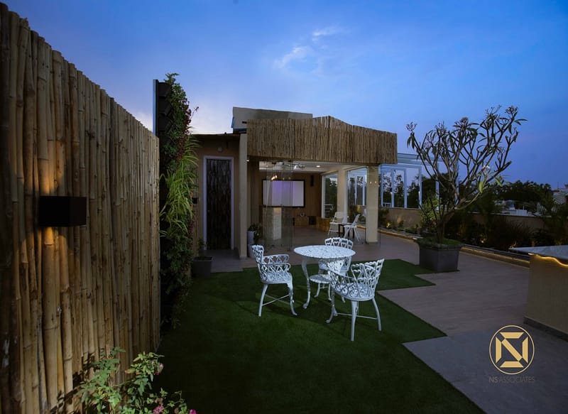 Evening rooftop garden with bamboo fencing, white wrought-iron table on green turf and planters, Hyderabad