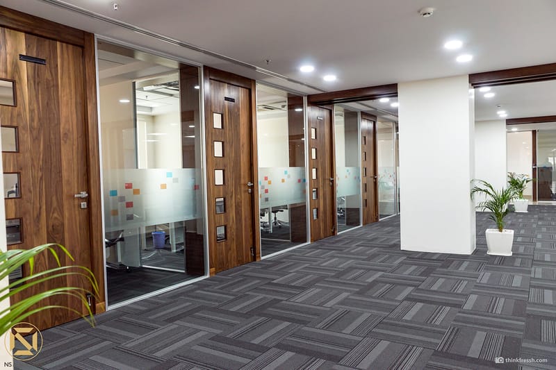 Office wing in Hyderabad with walnut-framed glass cabin doors, grey carpet tiles and white columns