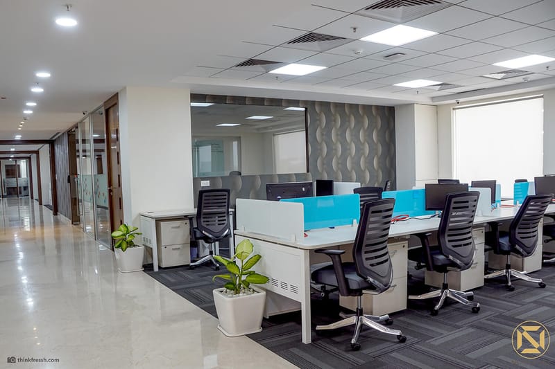 Open office bay in Hyderabad with white workstations, blue dividers, potted plants and a wave-textured accent wall