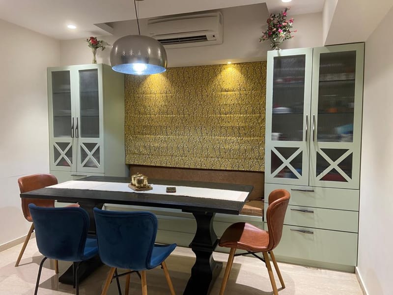 Dining area in Hyderabad with dark pedestal table, blue and tan velvet chairs and sage-green glass crockery cabinets