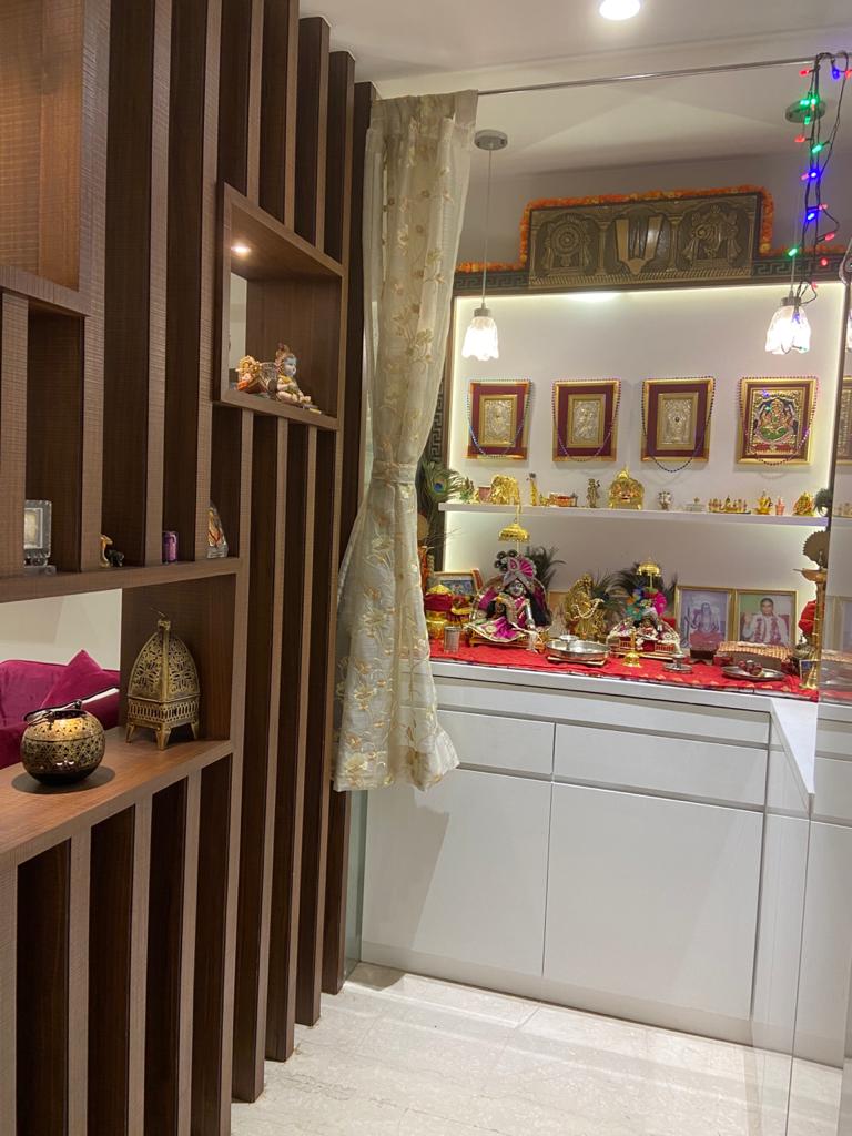 Home pooja unit with white cabinets, idols and framed deities, beside a dark wood-slat divider with lit niches, Hyderabad