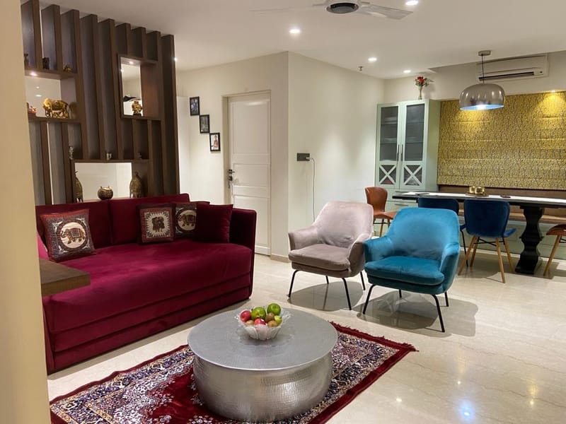 Living room with maroon velvet sofa, teal and grey accent chairs, silver drum coffee table and slatted wood divider, Hyderabad