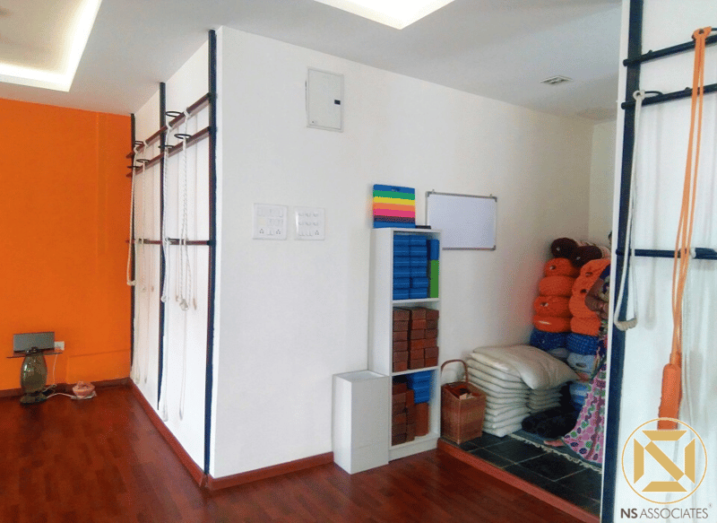Compact yoga prep room with bright orange accent wall, wall rope hooks, whiteboard and stacked bolsters, Hyderabad