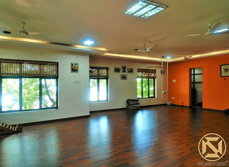 Open yoga practice hall with bright orange accent wall, glossy wood flooring and ceiling fans, Hyderabad