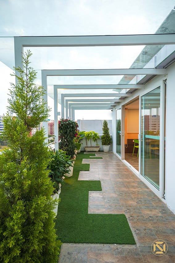Rooftop terrace walkway under a glass pergola, with tall conifers, an artificial-grass path and stone tiles beside glass doors, Hyderabad