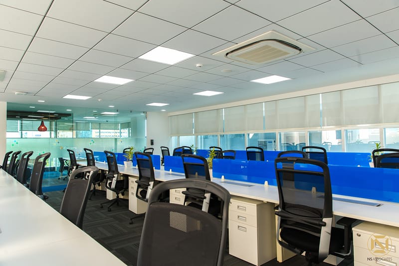 Office workstations with white desks, blue glass dividers, black mesh chairs and red pendant lamps under a grid ceiling, Hyderabad