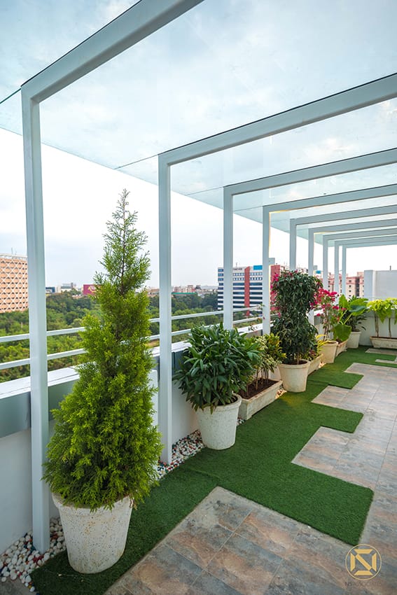Rooftop terrace with a steel-and-glass pergola, potted conifers and flowering plants over artificial grass and stone tiles, Hyderabad