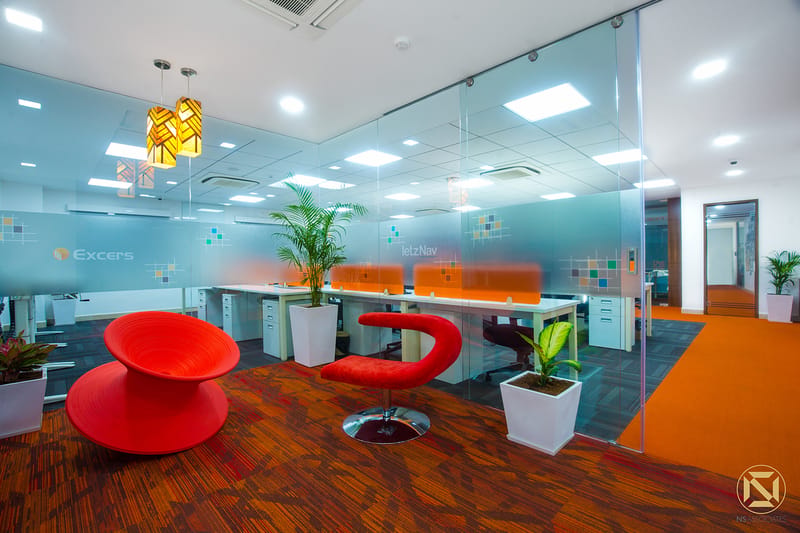 Office breakout zone with sculptural red chairs, yellow pendant lights and orange accents framed by glass partitions, Hyderabad