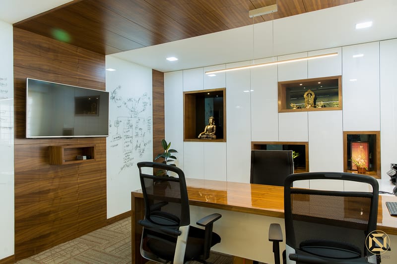 Office cabin with walnut wood paneling, wall-mounted TV, glossy white cabinets and lit display niches above a wood desk, Hyderabad