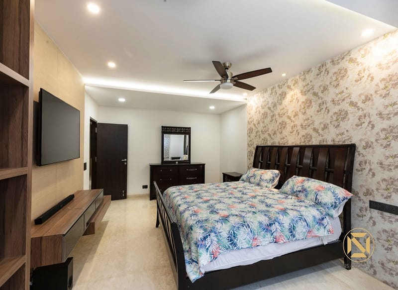 Bedroom in Hyderabad with dark-wood bed, tropical floral bedspread, vintage floral wallpaper, wall TV and open wood shelving