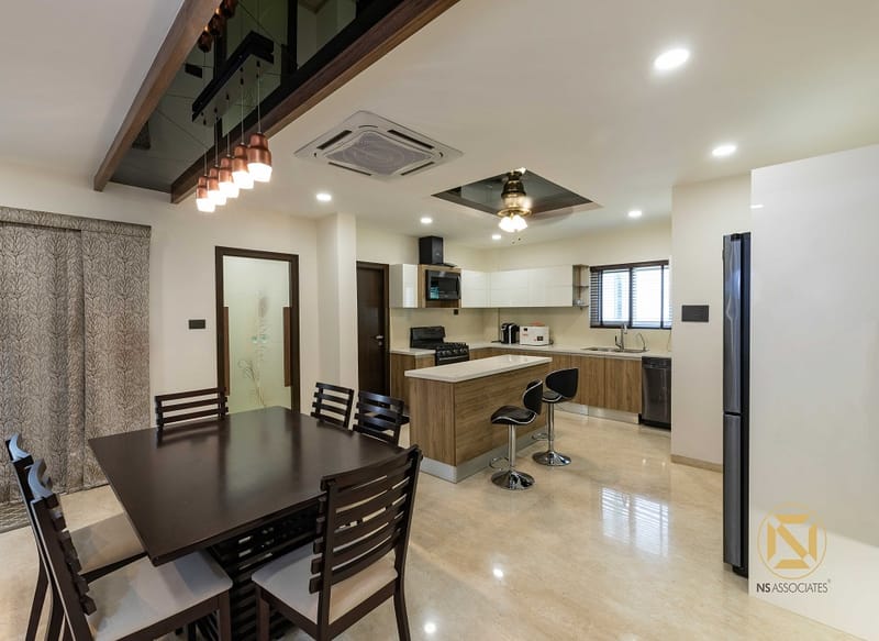 Dining area beside an L-shaped modular kitchen in Hyderabad, dark-wood table, bar stools and cluster pendant lights over the counter