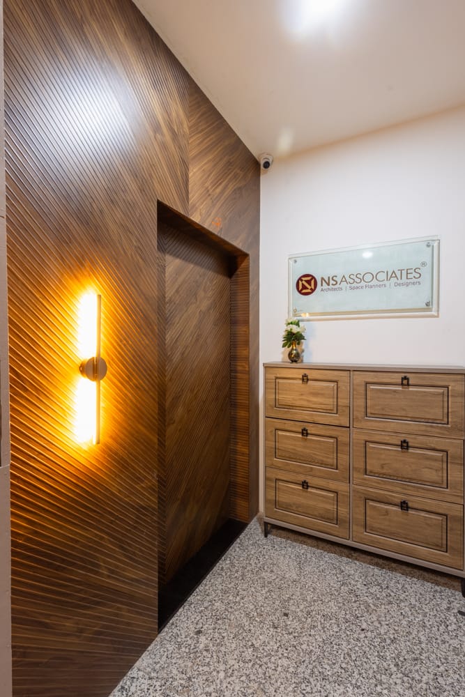 Office entry with fluted walnut wall panelling, a glowing linear wall light, terrazzo flooring and a wooden chest of drawers, Hyderabad