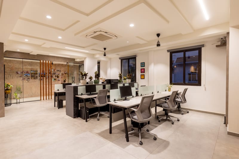 Open-plan office workstations with white desks, grey mesh chairs, coffered ceiling and cork feature wall behind glass, Hyderabad