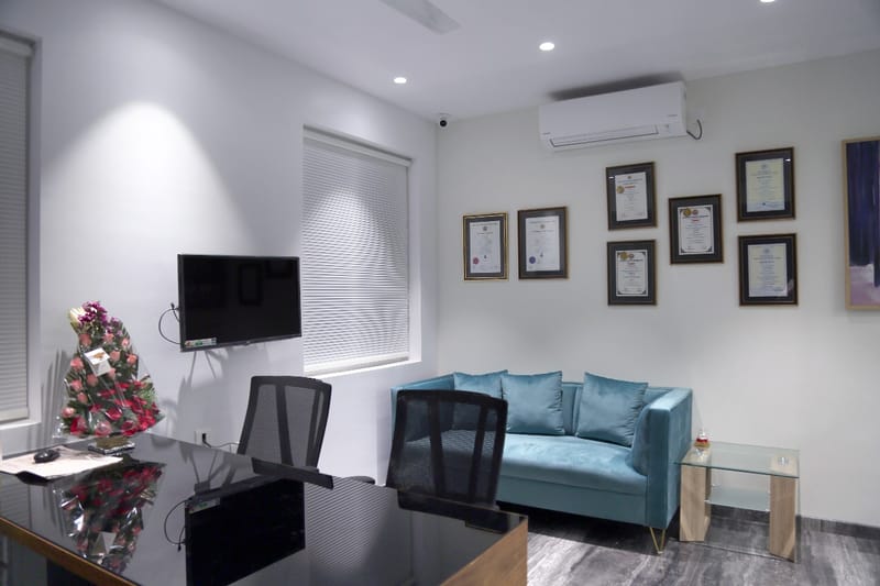 Compact cabin with glass desk, wall TV, teal sofa and a row of framed certificates, Hyderabad office