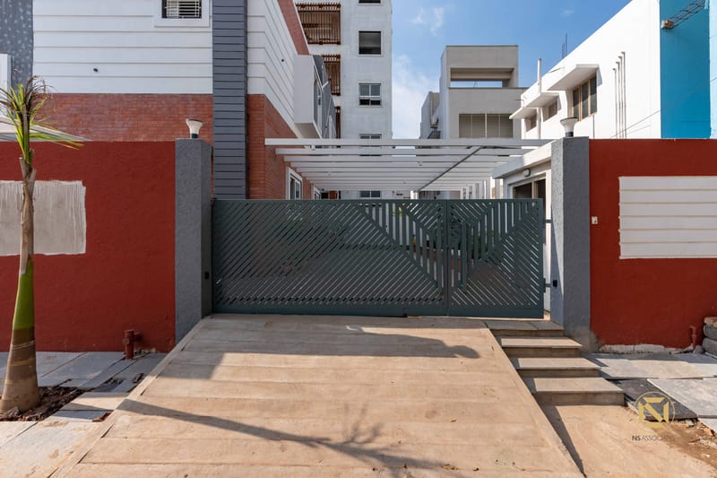 Street-front gateway of a Hyderabad home with angled green metal gate, red boundary wall and pergola-covered driveway