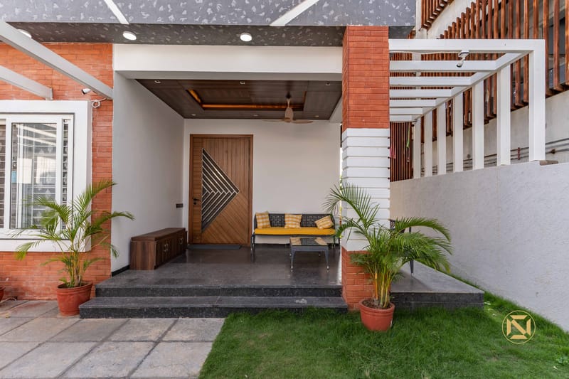 Home entrance porch with diamond-pattern wooden door, exposed brick column, slatted screen and a mustard cushioned bench, Hyderabad
