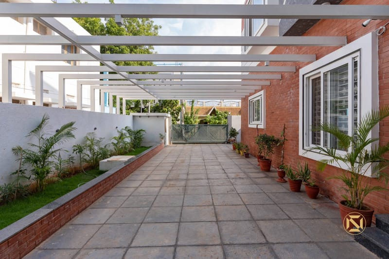 Home side passage with white steel pergola, exposed brick wall and potted palms lining tiled walkway, Hyderabad