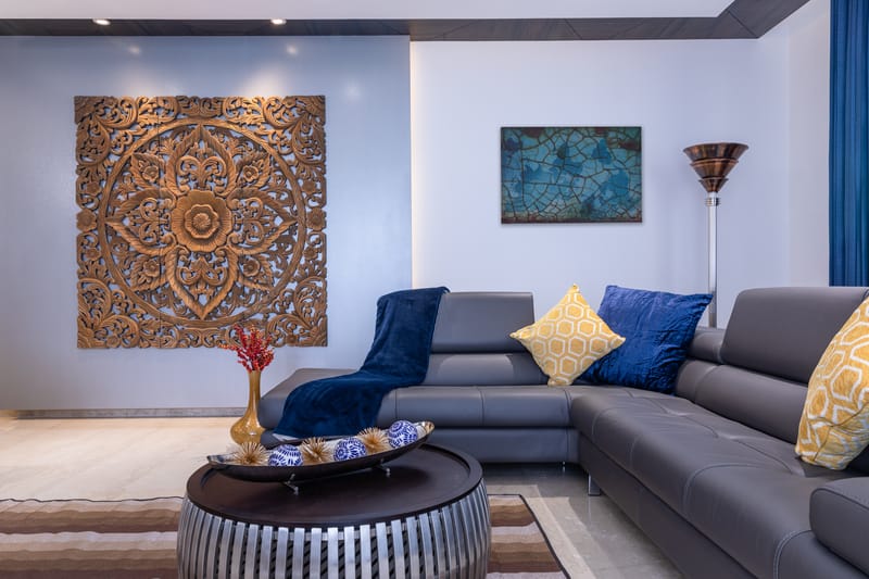 Carved wood mandala panel above a grey leather sectional with navy and yellow cushions and an arc floor lamp, Hyderabad living room