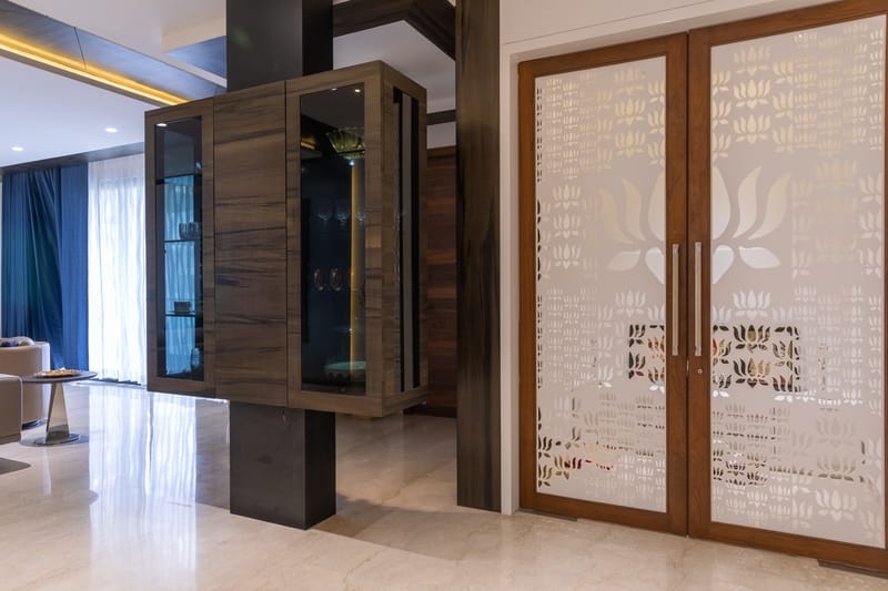 Wood-veneer display cabinet with backlit glass shelves beside lotus-motif laser-cut jali doors on a marble floor, Hyderabad