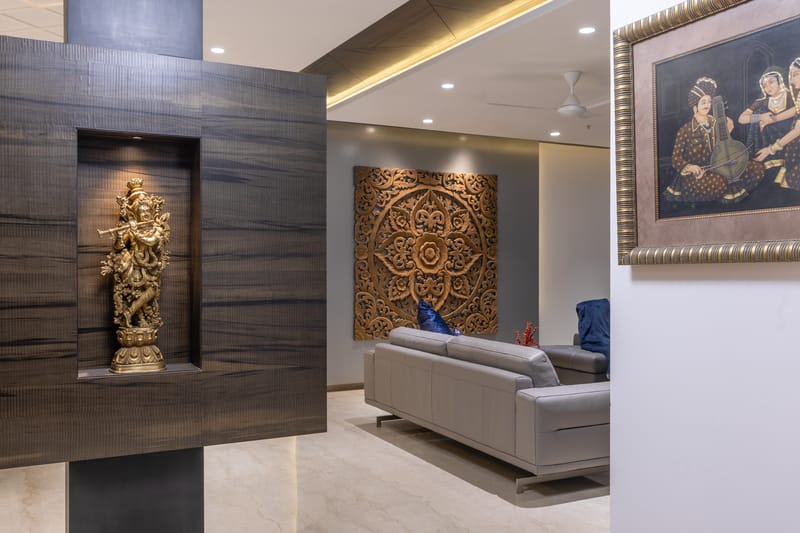 Living area with brass Krishna idol in a wood niche, large carved mandala panel, grey sofa and framed Indian art, Hyderabad