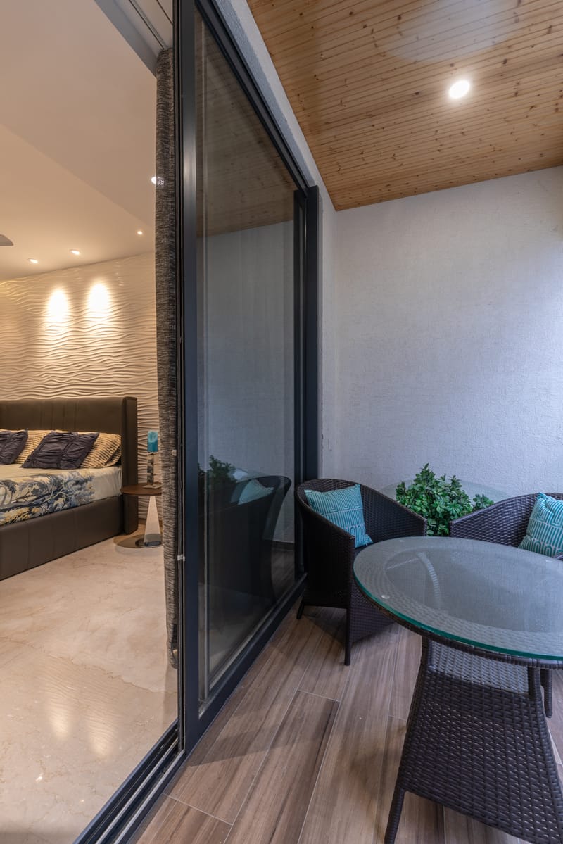 Balcony seating with woven chairs, glass table and wooden ceiling beside a wave-walled bedroom, Hyderabad apartment