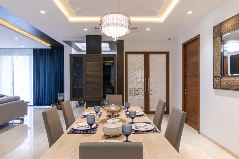 Dining room with light wood table for six, crystal pendant chandelier, navy drapes and a gilded mirror, Hyderabad home
