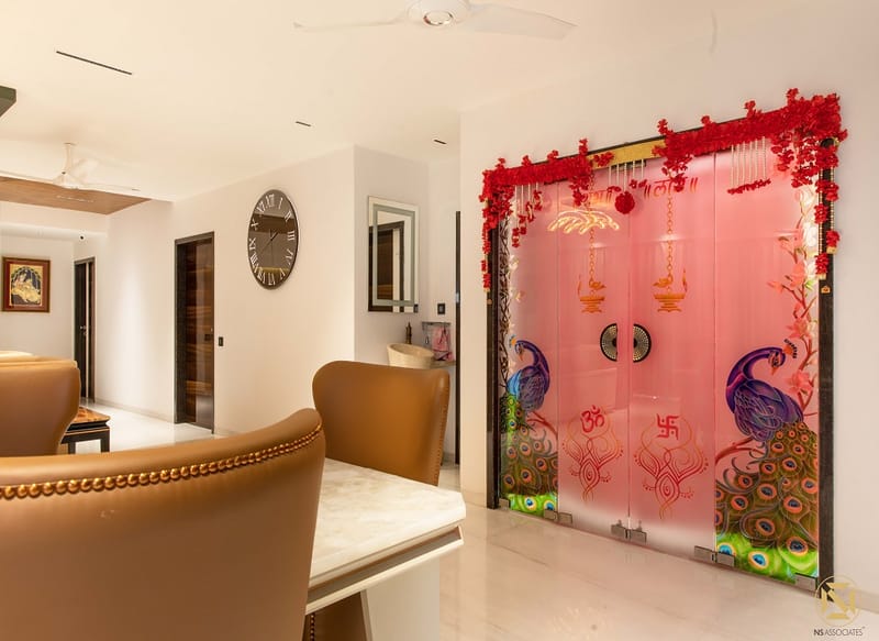 Pooja room with backlit peacock-motif glass doors framed by marigold garlands beside a marble dining nook, Hyderabad