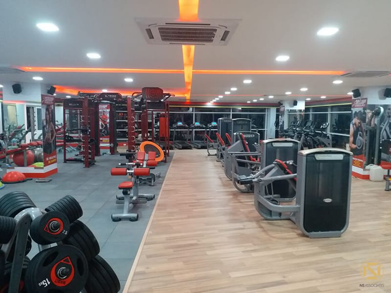 Wide gym view with dumbbell rack, leg-press machines and orange cove lighting along wood flooring, Hyderabad
