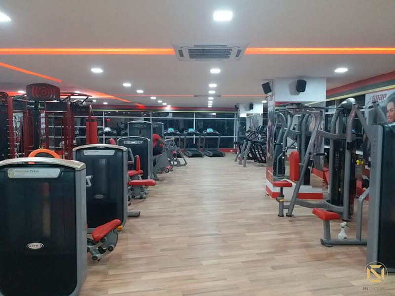Gym interior with rows of strength equipment, red benches and mirrored walls along a wood-floor aisle, Hyderabad