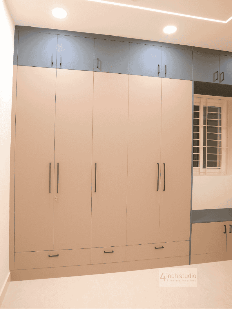 Large beige wardrobe with navy upper loft cabinets and lower drawers beside a window-seat bench, Hyderabad bedroom