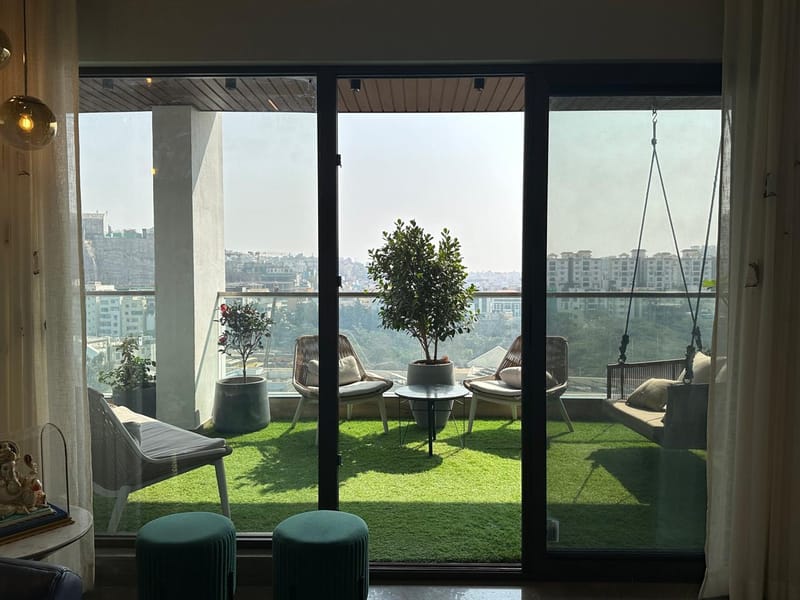 High-rise balcony with artificial-turf floor, woven lounge chairs and potted tree framed by glass doors, Hyderabad