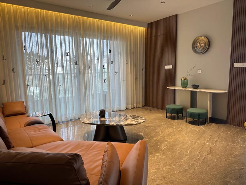 Living room with tan leather sofas, sheer floor-to-ceiling drapes, wood-slat wall and marble console with green stools, Hyderabad