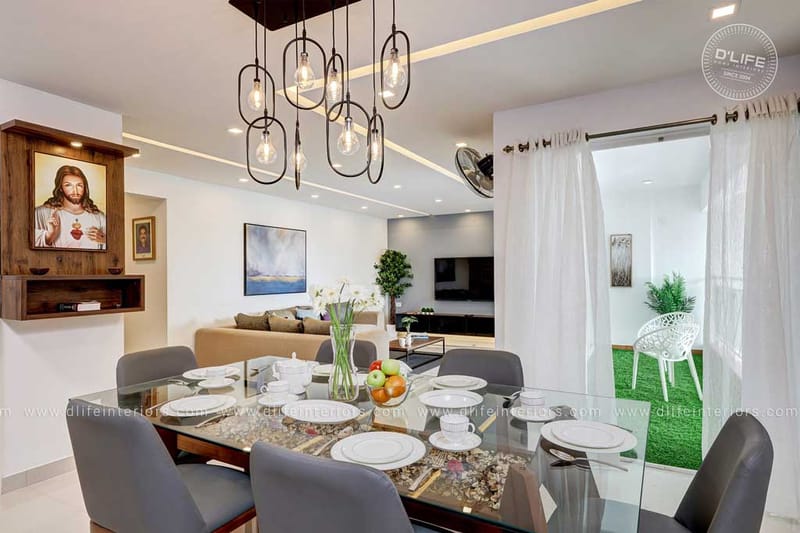 Open-plan dining and living area with glass table, geometric pendants and a wood Sacred Heart shrine niche, Hyderabad home