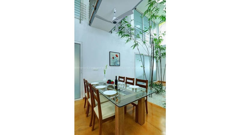 Glass-top dining table with timber chairs beside a tall bamboo planter in a double-height atrium, Hyderabad