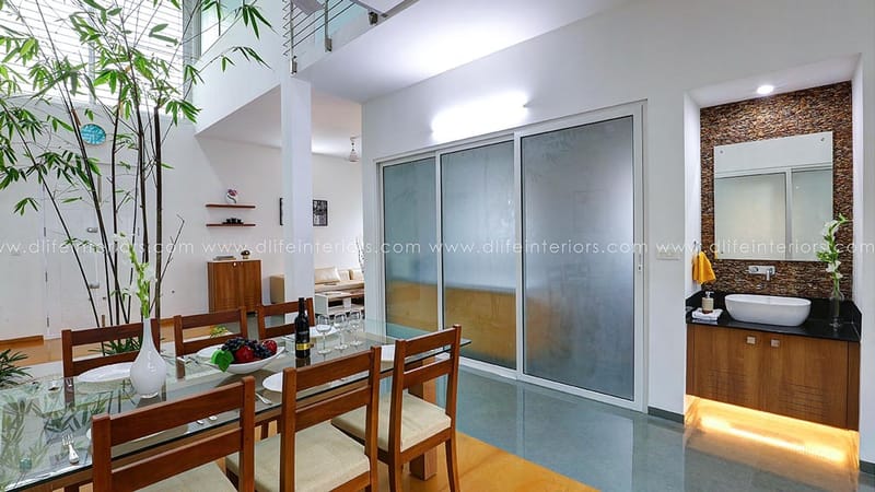Double-height dining area in Hyderabad with glass-top wooden table, bamboo plant and mosaic-tiled washbasin niche