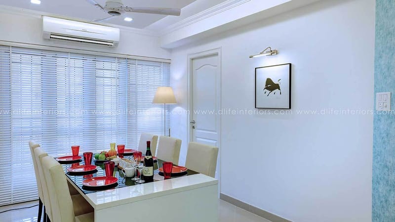 Bright white dining area with glass-top table, red and white place settings, framed art and window blinds, Hyderabad home