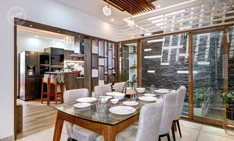 Dining area with glass-top wooden table, grey tufted chairs and stone-clad wall beside open kitchen, Hyderabad