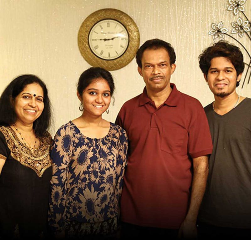 Vijayakumar family portrait of four beside a wall clock and metal floral wall art in their Hyderabad home