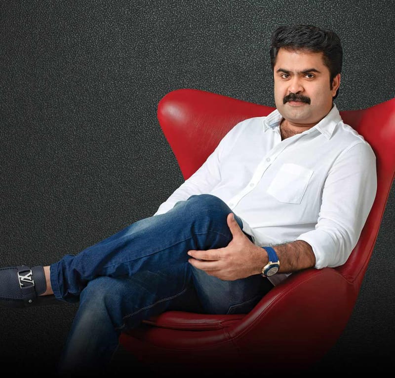 Portrait of a man in white shirt and jeans seated in a red leather wingback chair against a textured dark wall
