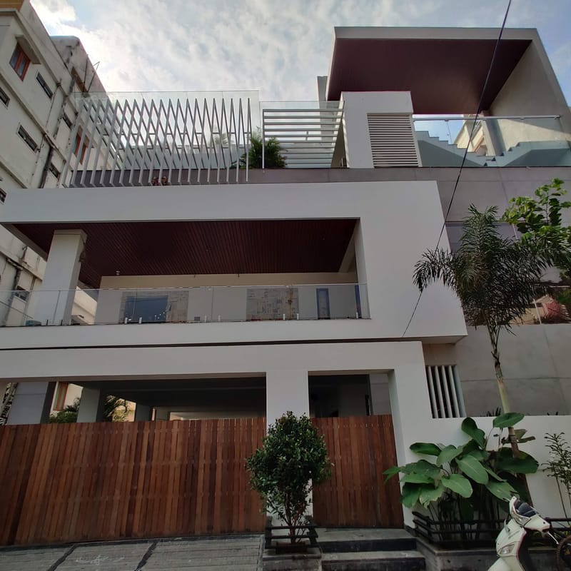 Modern white-and-grey house front with wood louvers, glass balcony railings and a slatted rooftop screen in Hyderabad