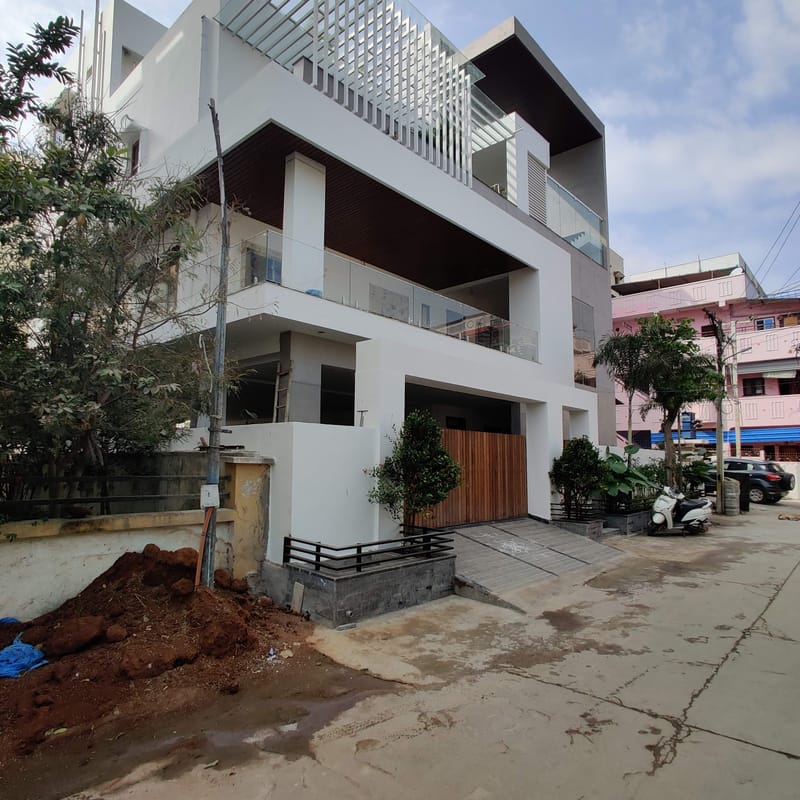 Street-side view of a white contemporary house with timber-clad gate, glass balustrades and rooftop louvres, Hyderabad