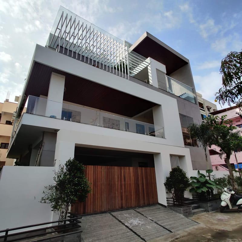 Three-storey modern house with white render, timber garage door, glass balconies and rooftop louvered screen, Hyderabad
