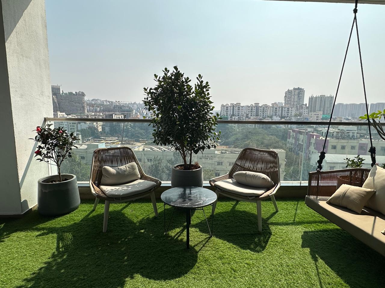 Open balcony with green turf, woven armchairs, potted tree and glass railing over city skyline, Hyderabad