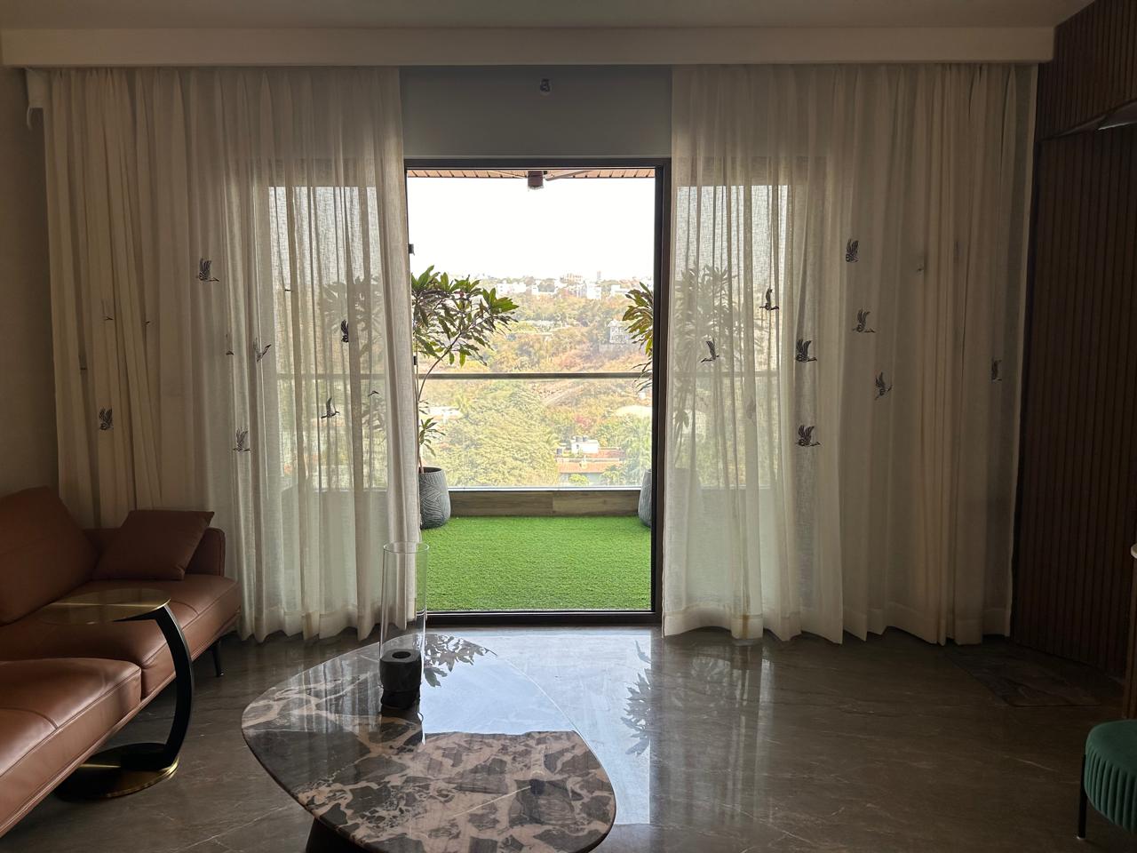 Living room balcony view through sheer white curtains to artificial-turf balcony and city greenery, marble floor, Hyderabad