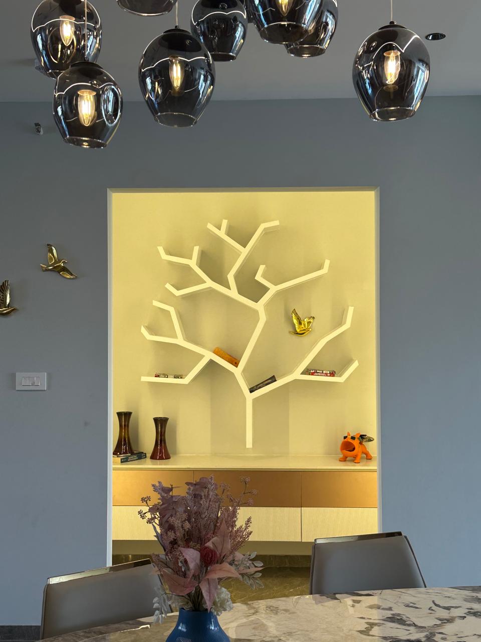 Dining area featuring a sculptural tree bookshelf in a yellow alcove, chrome globe pendant cluster and marble table, Hyderabad