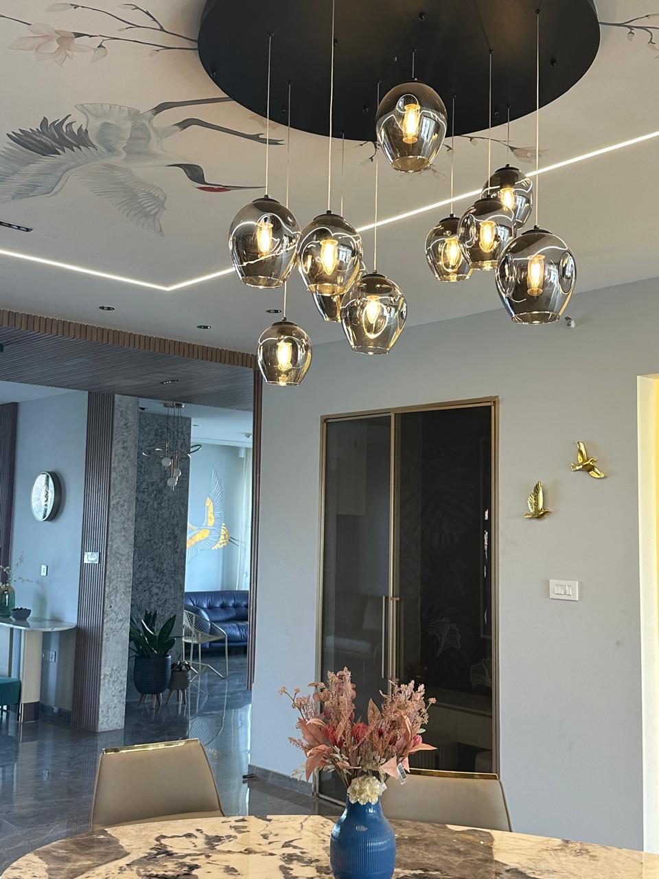 Dining area looking up at a chrome globe pendant cluster on a black ceiling disc with crane ceiling mural and brass birds, Hyderabad